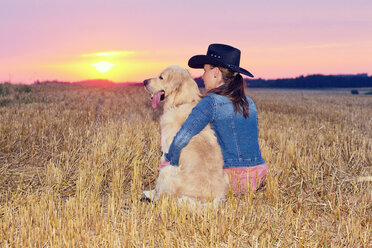 Junge Frau mit Golden Retriever sitzt in der Dämmerung auf einem Stoppelfeld - MAEF010833