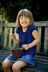 Grinsendes kleines Mädchen, das auf einer Gartenbank mit einem Weidenkorb voller Blaubeeren sitzt - LVF003736