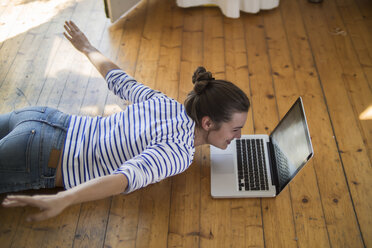 Young woman at home lying on wooden floor with laptop, pretending to fly - RIBF000182