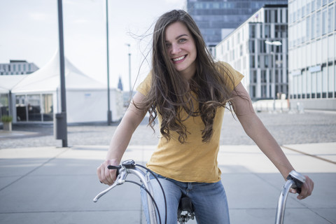 Deutschland, Köln, Porträt einer lächelnden jungen Frau mit wehendem Haar auf ihrem Fahrrad, lizenzfreies Stockfoto