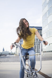 Germany, Cologne, portrait of smiling young woman with blowing hair on her bicycle - RIBF000215