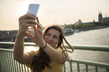 Germany, Cologne, young woman standing on Rhine bridge taking a selfie with her smartphone - RIBF000230