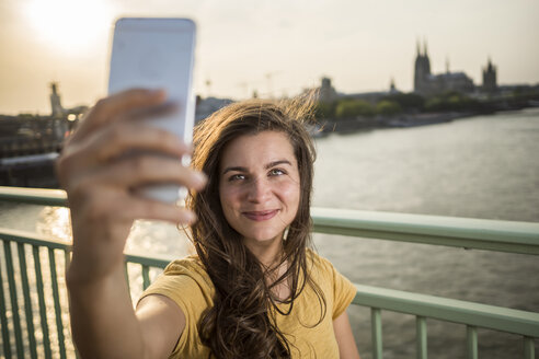 Germany, Cologne, young woman standing on Rhine bridge taking a selfie with her smartphone - RIBF000231