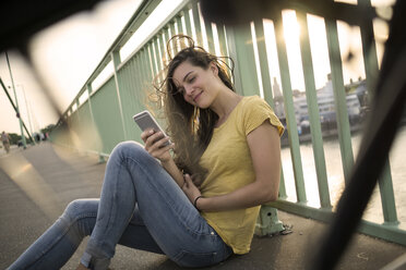 Deutschland, Köln, Frau sitzt auf der Rheinbrücke und schaut auf ihr Smartphone - RIBF000232