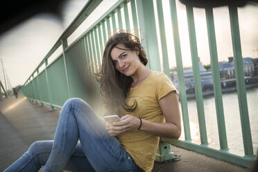 Germany, Cologne, woman sitting on Rhine bridge looking at her smartphone - RIBF000233