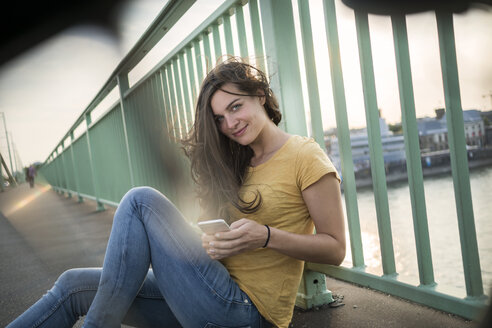 Deutschland, Köln, Frau sitzt auf der Rheinbrücke und schaut auf ihr Smartphone - RIBF000233