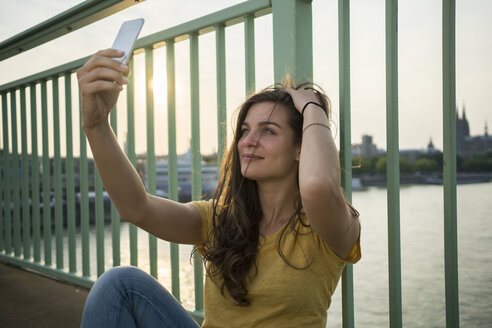 Deutschland, Köln, junge Frau sitzt auf der Rheinbrücke und macht ein Selfie mit ihrem Smartphone - RIBF000234