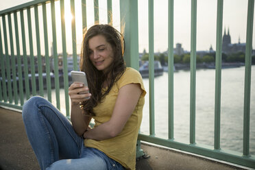 Deutschland, Köln, junge Frau sitzt auf der Rheinbrücke und schaut auf ihr Smartphone - RIBF000235