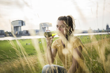 Germany, Cologne, young woman drinking beer out of a bottle on a meadow near Rhine River - RIBF000223