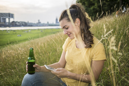 Deutschland, Köln, Frau mit Bierflasche und Smartphone entspannt auf einer Wiese am Rhein - RIBF000224