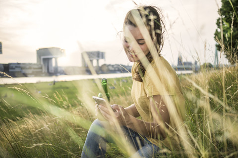 Germany, Cologne, woman with beer bottle and smartphone relaxing on a meadow near Rhine River stock photo