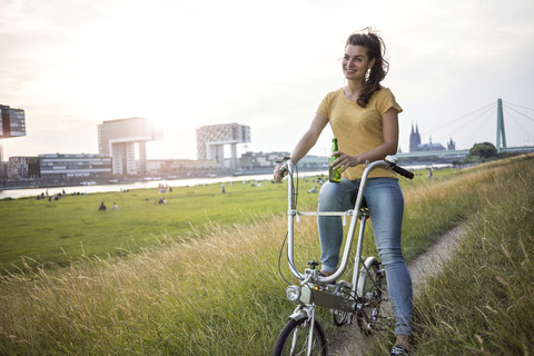 Deutschland, Köln, junge Frau mit Fahrrad auf Wiese am Rhein in der Abenddämmerung, lizenzfreies Stockfoto