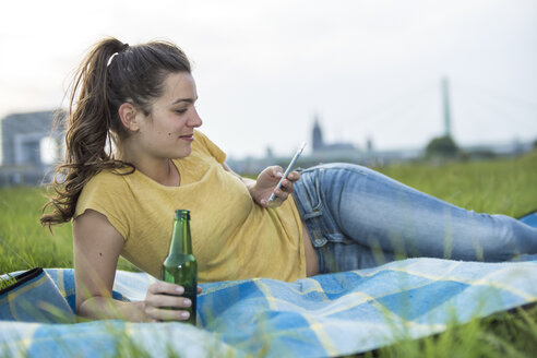 Deutschland, Köln, Frau mit Bierflasche und Smartphone entspannt auf einer Wiese am Rhein - RIBF000228