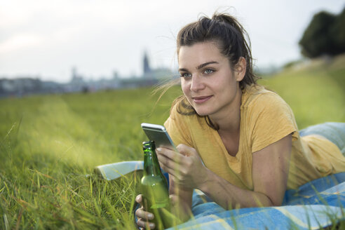 Deutschland, Köln, Frau mit Bierflasche und Smartphone entspannt auf einer Wiese in der Abenddämmerung - RIBF000245