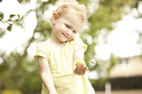 Kleines Mädchen mit Apfel in der Hand im Garten stehend, lizenzfreies Stockfoto