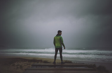 Spanien, Valdovino, Rückansicht eines Joggers, der an einem stürmischen Tag am Meer steht - RAEF000270