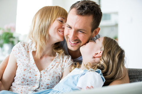 Glückliche Familie sitzt auf der Couch, Mutter und Tochter küssen den Vater, lizenzfreies Stockfoto