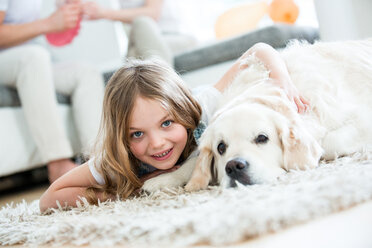 Kleines Mädchen kuschelt mit ihrem Hund, auf dem Boden liegend, Eltern im Hintergrund - WESTF021546