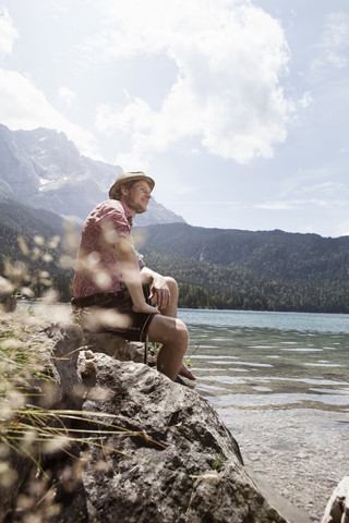 Germany, Bavaria, Eibsee, smiling man in lederhosen sitting on lakeshore stock photo