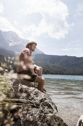 Deutschland, Bayern, Eibsee, lächelnder Mann in Lederhosen am Seeufer sitzend - RBF003008