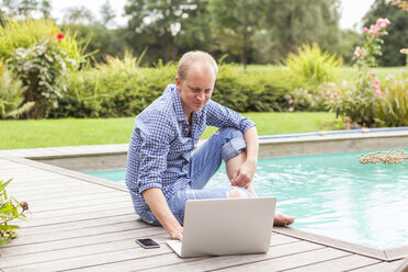 Man sitting on floor beside a pool using laptop - JUNF000396