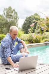 Man sitting on floor beside a pool using laptop - JUNF000397