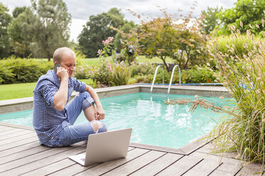 Man with laptop sitting beside a pool telephoning with smartphone - JUNF000398