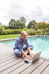 Mann sitzt auf dem Boden neben einem Pool und benutzt einen Laptop - JUNF000400