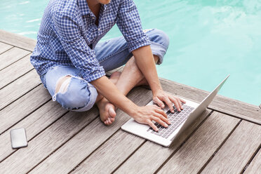 Mann sitzt auf Holzboden neben einem Pool und benutzt einen Laptop - JUNF000387