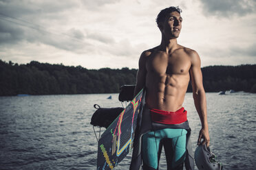 Young wakeboarder standing with his equipment at lakeshore - MADF000504