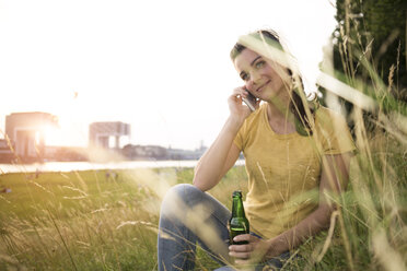 Deutschland, Köln, junge Frau mit Bierflasche, die auf einer Wiese am Rhein sitzt und telefoniert - RIBF000146