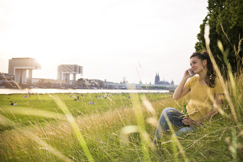 Germany, Cologne, young woman with beer bottle sitting on a meadow near Rhine River telephoning stock photo