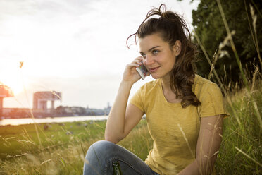Germany, Cologne, young woman sitting on a meadow near Rhine River telephoning - RIBF000144