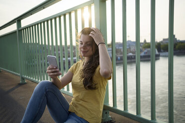 Germany, Cologne, young woman with smartphone sitting on Rhine Bridge - RIBF000147