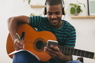 Mann mit Gitarre und Kopfhörern schaut auf sein Smartphone und schreibt etwas auf - EBSF000856