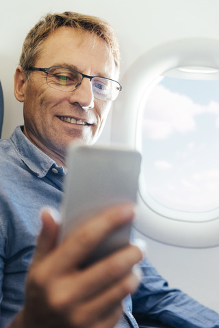 Porträt eines lächelnden Mannes, der in einem Flugzeug sitzt und auf sein Smartphone schaut, lizenzfreies Stockfoto