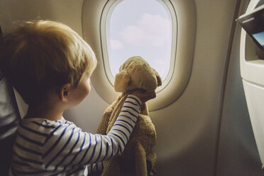 Kleiner Junge sitzt mit seinem Kuscheltier in einem Flugzeug und schaut durch das Fenster - MFF001990