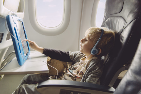 Kleiner Junge sitzt in einem Flugzeug und sieht sich etwas auf einem digitalen Tablet an, lizenzfreies Stockfoto