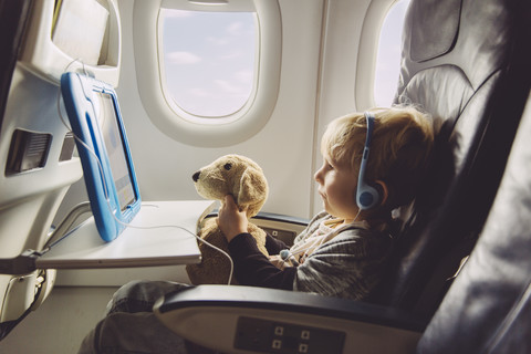 Kleiner Junge sitzt in einem Flugzeug und sieht sich etwas auf einem digitalen Tablet an, lizenzfreies Stockfoto