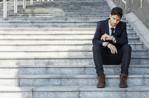 Porträt eines jungen Geschäftsmannes, der auf einer Treppe sitzt und die Zeit kontrolliert - PASF000007