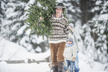 Österreich, Altenmarkt-Zauchensee, fröhlicher Mann trägt Weihnachtsbaum auf der Schulter im Winterwald - HHF005383