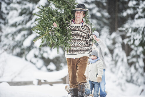 Austria, Altenmarkt-Zauchensee, happy man carrying Christmas tree on his shoulder in winter forest - HHF005383
