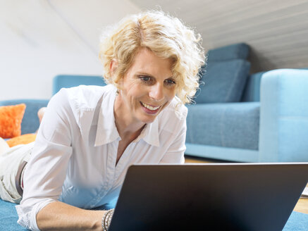 Porträt einer lächelnden blonden Frau, die auf dem Boden liegt und einen Laptop benutzt - LAF001461