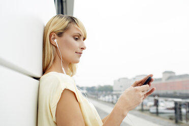 Blonde Frau lehnt an der Fassade und hört Musik mit MP3-Player und Kopfhörern - PESF000035