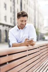 Porträt eines lächelnden Mannes, der auf einer Holzbank sitzt und auf sein Smartphone schaut - PESF000039