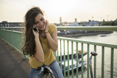Junge Frau mit Fahrrad auf der Rheinbrücke stehend und mit Smartphone telefonierend - RIBF000243