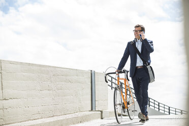 Businessman pushing bike while talking on the phone - UUF005330
