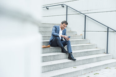 Businessman sitting on stairs using smart phone - UUF005219
