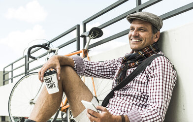 Mature man with bicycle leaning on wall, holding coffee cup and smart phone - UUF005256