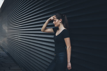 Junge Frau mit Hand auf dem Kopf vor einer schwarzen Fassade stehend - CHAF001224